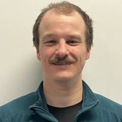 Middle-aged man with mustache wearing a dark teal jacket over dark shirt, smiling at camera against neutral background