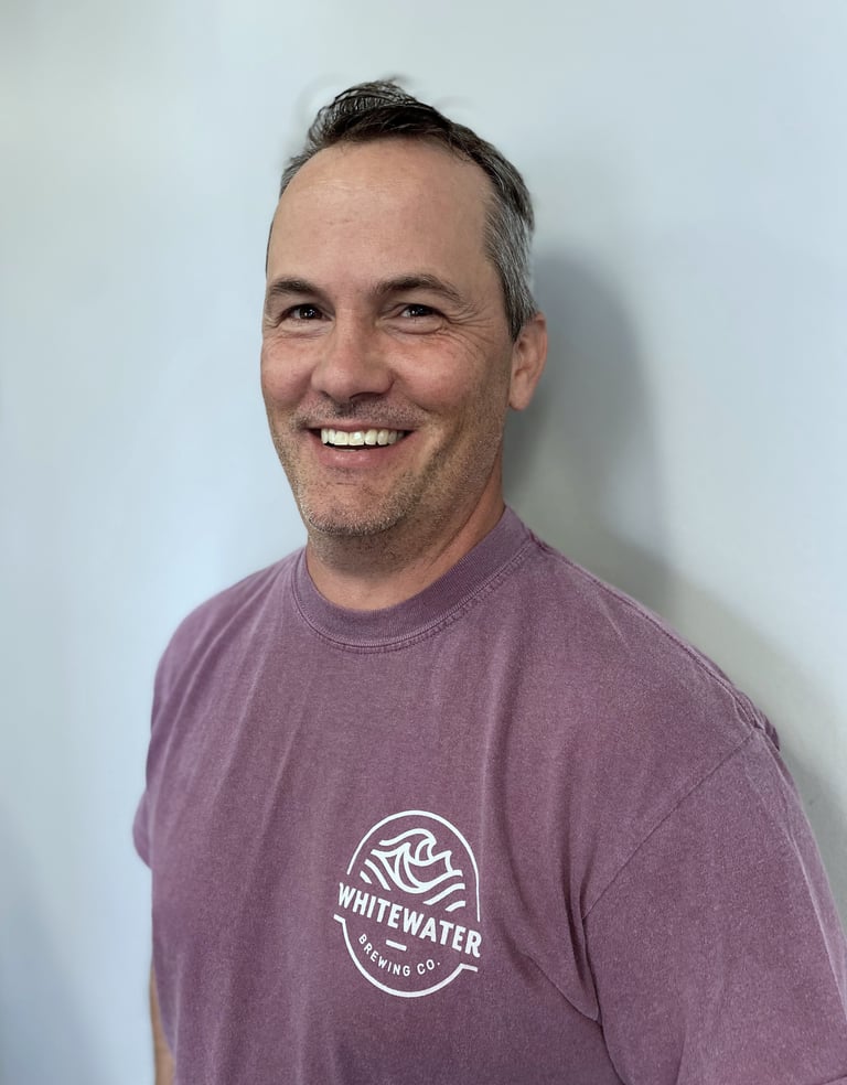 Smiling man wearing a purple Whitewater shirt against a light gray background, headshot photo