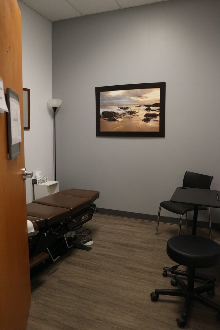 Medical examination room with brown treatment table, adjustable stool, framed beach landscape artwork, and minimal furniture on wooden flooring