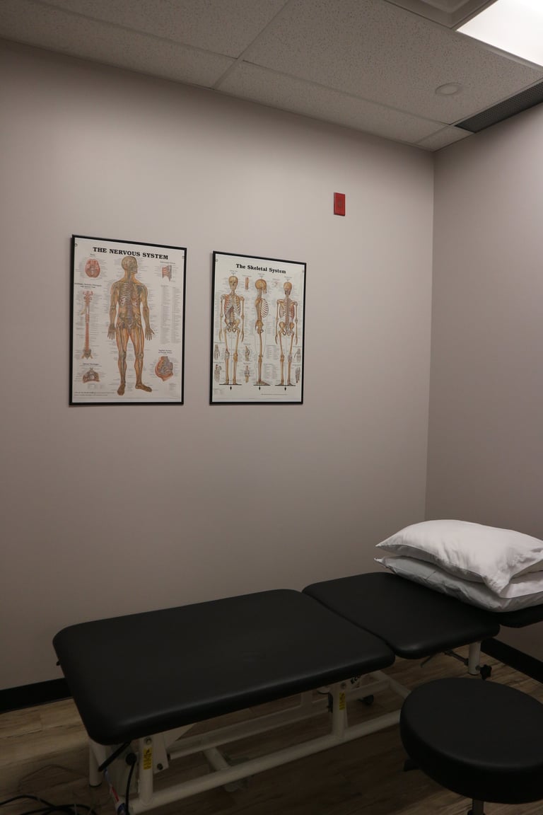 Medical examination room with anatomical posters of the nervous and skeletal systems on white walls, and a black treatment table with white pillow