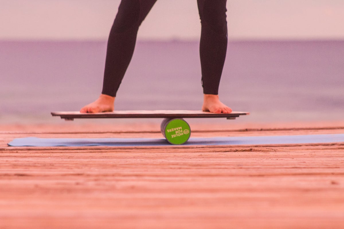 Person balancing on wooden board outdoors with a green fitness ball underneath, legs visible in dark tights and orange shoes