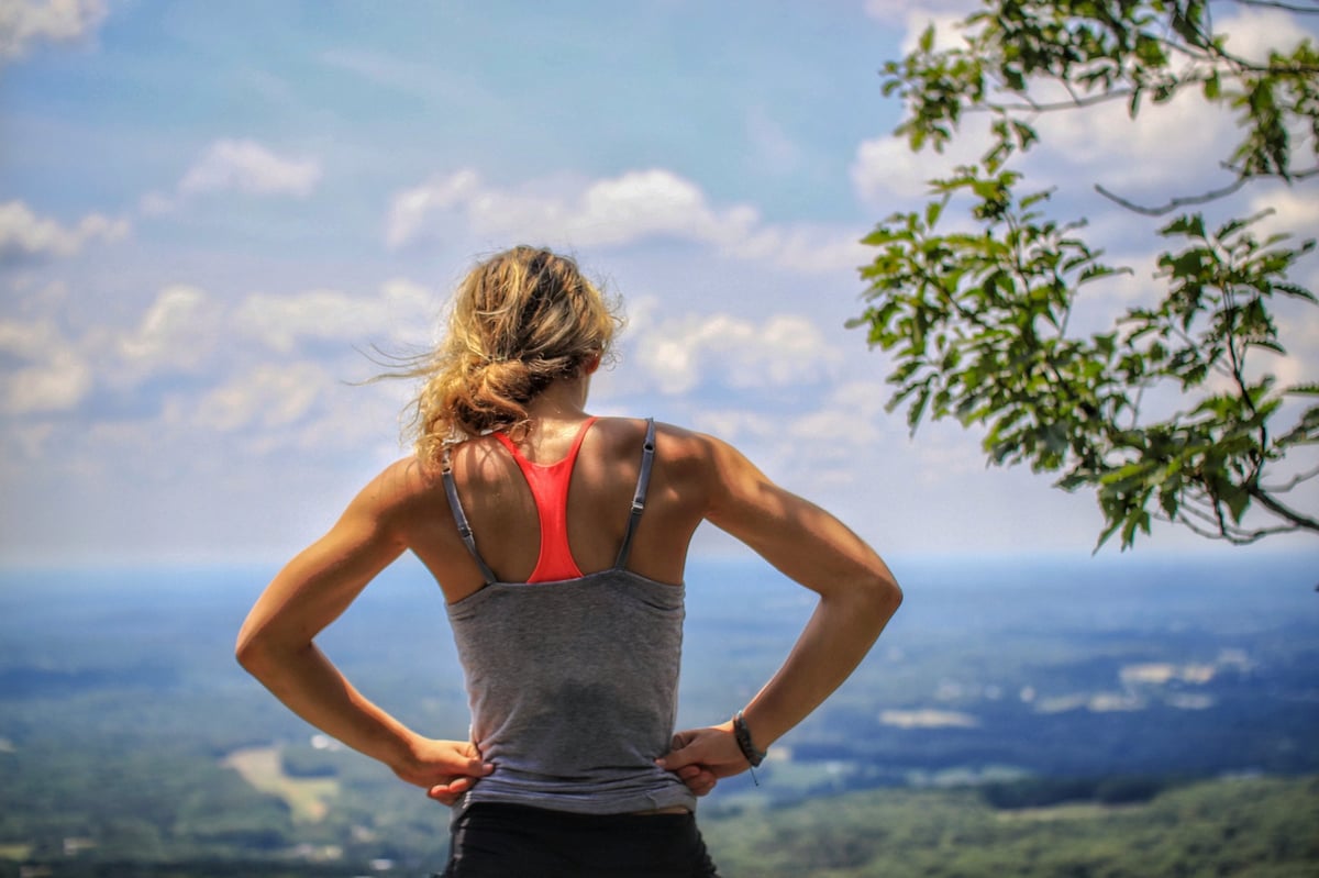 Woman in athletic wear standing on mountain overlook with hands on hips, viewing expansive landscape below