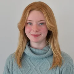 Portrait of a young woman with long reddish-brown hair wearing a gray turtleneck sweater against a white background