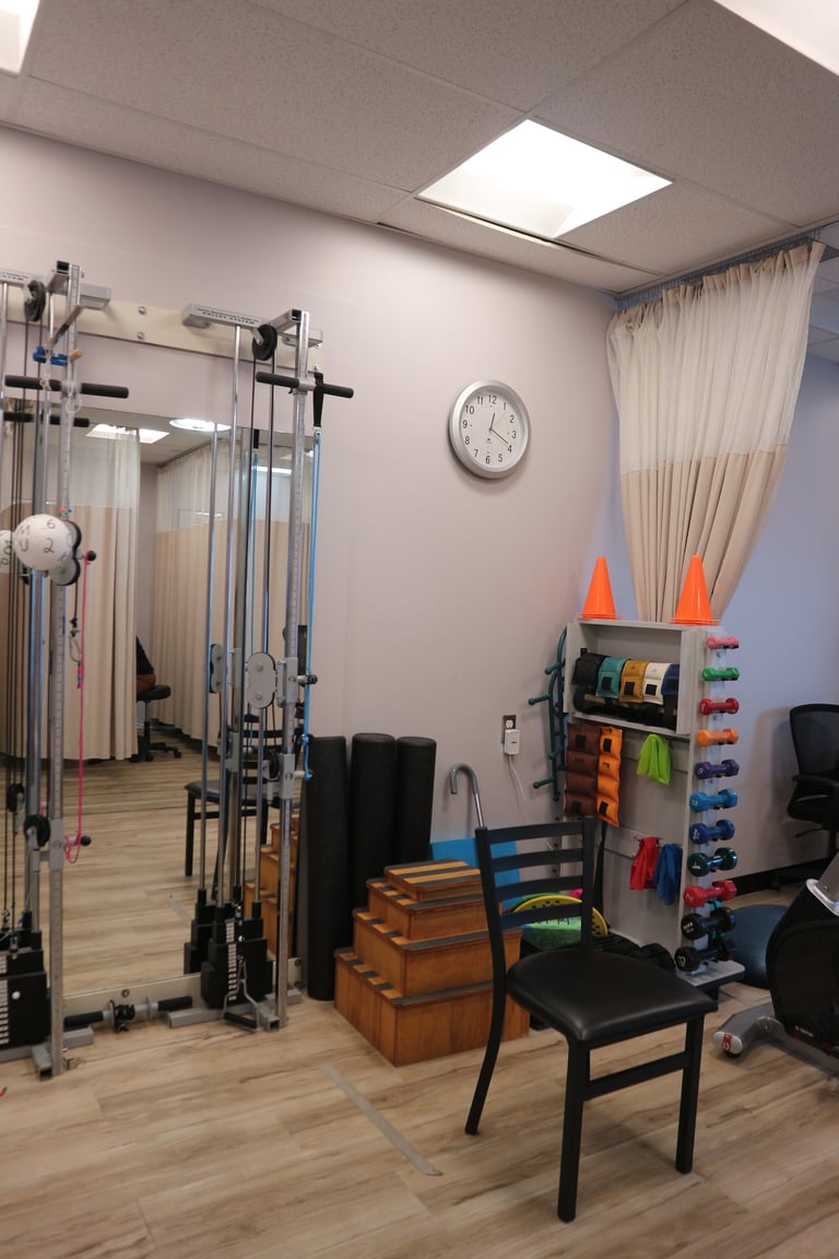 Physical therapy clinic room with exercise equipment, resistance bands on wall rack, cable machine, wooden step platform, and black chair