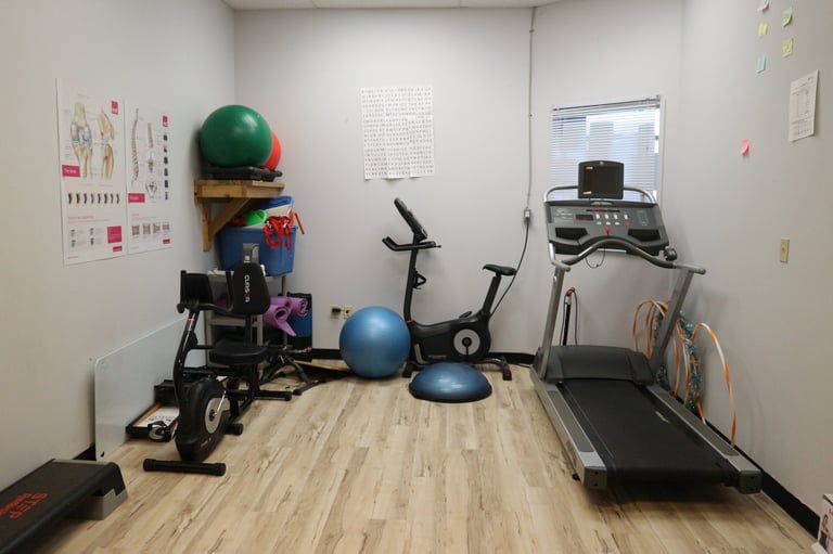 Home gym room with exercise equipment including treadmill, stationary bike, bosu ball, fitness balls, and workout posters on white walls