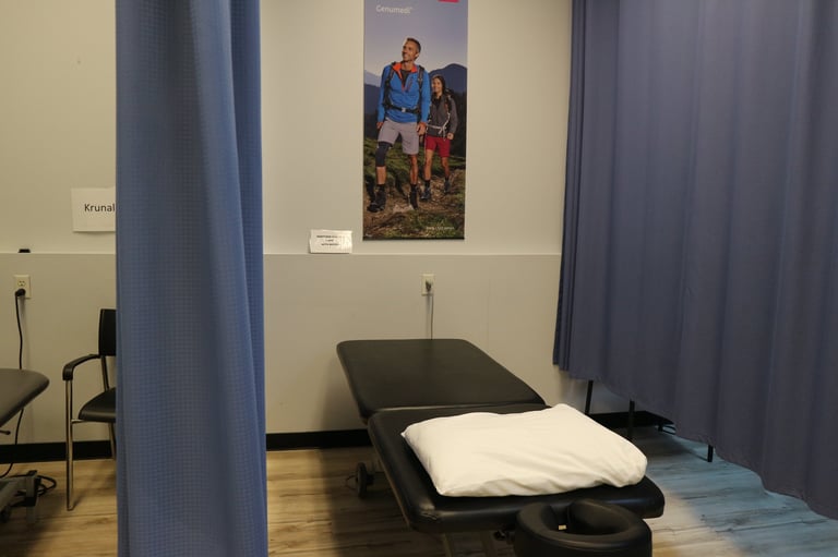 Medical examination room with black treatment table, white pillow, blue curtains, wooden floor, and family photo on wall
